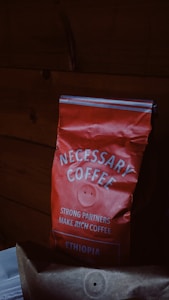 A red bag of coffee labeled 'NECESSARY COFFEE' with additional text stating 'STRONG PARTNERS MAKE RICH COFFEE' and 'ETHIOPIA' on it. The bag appears slightly crumpled and is set against a dark wooden background.