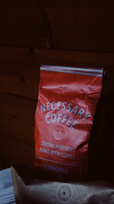 A red bag of coffee labeled 'NECESSARY COFFEE' with additional text stating 'STRONG PARTNERS MAKE RICH COFFEE' and 'ETHIOPIA' on it. The bag appears slightly crumpled and is set against a dark wooden background.