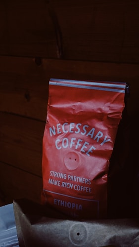 A red bag of coffee labeled 'NECESSARY COFFEE' with additional text stating 'STRONG PARTNERS MAKE RICH COFFEE' and 'ETHIOPIA' on it. The bag appears slightly crumpled and is set against a dark wooden background.