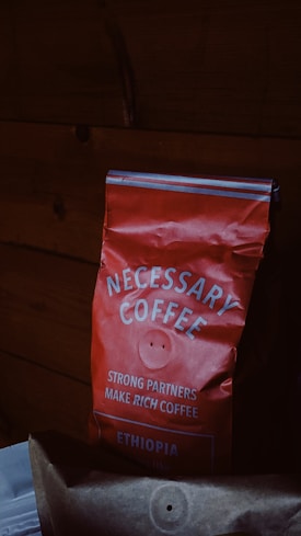 A red bag of coffee labeled 'NECESSARY COFFEE' with additional text stating 'STRONG PARTNERS MAKE RICH COFFEE' and 'ETHIOPIA' on it. The bag appears slightly crumpled and is set against a dark wooden background.