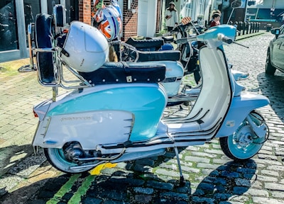 A classic scooter with a blue and white color scheme is parked on a cobblestone street. A helmet with a Union Jack design rests on the scooter's back seat. In the background, two people are walking near a brick building with large glass windows. Another scooter is parked behind, partially visible.