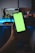 A hand is holding a smartphone with a bright green screen, in front of a desk setup featuring a dual monitor display. The monitors show scenes from games, with one depicting a driving game and the other an action game. The background is dimly lit, creating a contrast with the screen's brightness.