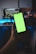 A hand is holding a smartphone with a bright green screen, in front of a desk setup featuring a dual monitor display. The monitors show scenes from games, with one depicting a driving game and the other an action game. The background is dimly lit, creating a contrast with the screen's brightness.