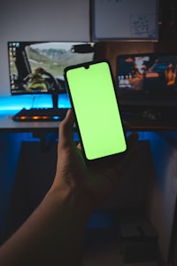 A hand is holding a smartphone with a bright green screen, in front of a desk setup featuring a dual monitor display. The monitors show scenes from games, with one depicting a driving game and the other an action game. The background is dimly lit, creating a contrast with the screen