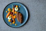 A beautifully arranged gourmet dish on a blue ceramic plate, featuring slices of roast meat and an artistic assortment of colorful vegetables including carrots, onions, and cherries. Small dots of vibrant orange puree add a touch of elegance.