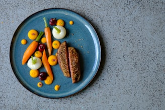 A beautifully arranged gourmet dish on a blue ceramic plate, featuring slices of roast meat and an artistic assortment of colorful vegetables including carrots, onions, and cherries. Small dots of vibrant orange puree add a touch of elegance.