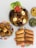 A variety of dishes are displayed, including a bowl of jollof rice garnished with boiled eggs, grilled chicken, and vegetables, a plate of spring rolls, a bowl of golden puff-puff, a dish with green vegetables, and a bowl of beans porridge. The setup highlights vibrant colors and textures.