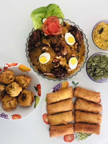 A variety of dishes are displayed, including a bowl of jollof rice garnished with boiled eggs, grilled chicken, and vegetables, a plate of spring rolls, a bowl of golden puff-puff, a dish with green vegetables, and a bowl of beans porridge. The setup highlights vibrant colors and textures.