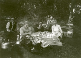 A group of five people sitting on a blanket in a wooded outdoor setting, enjoying a picnic. They are surrounded by picnic items such as a kettle, bottles, and food containers. The people appear to be in a relaxed and jovial mood, with some smiling and conversing with each other.