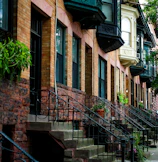 Brick row houses with ornate black iron railings line a quaint street. The buildings feature a mix of architectural styles, with large windows and decorative cornices. Lush greenery and potted plants add a touch of nature to the urban setting.