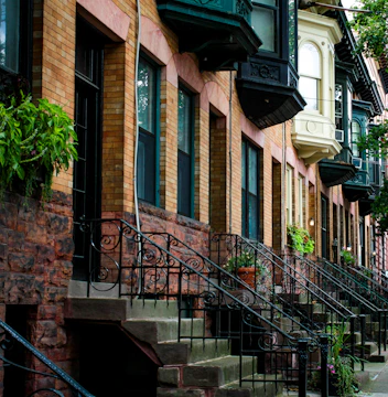 Brick row houses with ornate black iron railings line a quaint street. The buildings feature a mix of architectural styles, with large windows and decorative cornices. Lush greenery and potted plants add a touch of nature to the urban setting.