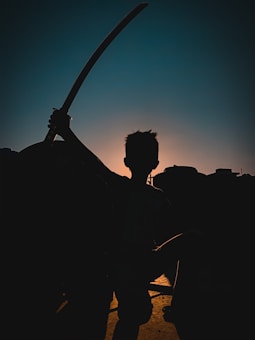A silhouetted figure of a person holding a sword against a backdrop of a sunrise or sunset. The figure stands prominently with dramatic lighting creating a contrast between the dark foreground and the vibrant sky.