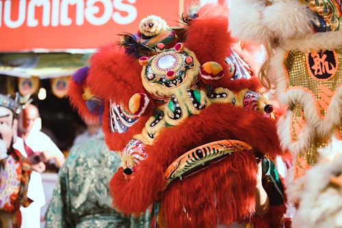 A vibrant and colorful scene featuring traditional Chinese lion dance costumes with intricate embroidery and fluffy textures. The setting seems lively and festive with multiple people wearing detailed headpieces and elaborate garments. The focus is on the red and gold lion costume adorned with feathers and ornate designs, indicating a cultural celebration.