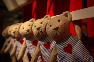 A row of identical plush teddy bears hanging side by side, each wearing a blue and white striped outfit adorned with a red heart on their chest. The background is predominantly red, enhancing the warm ambiance.