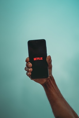 A hand holding a smartphone with the Netflix logo displayed on the screen against a light blue background.