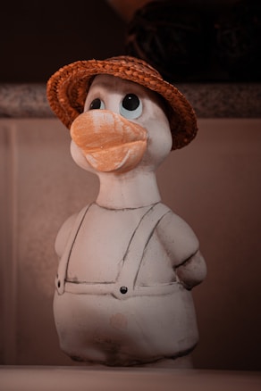 A ceramic duck figurine is dressed in overalls and sporting a woven straw hat. The figure has a large orange beak and expressive eyes, situated on a plain background with muted colors.