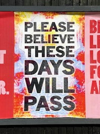 A large sign with the message 'PLEASE BELIEVE THESE DAYS WILL PASS' displayed against a colorful background that transitions from yellow to blue to red. The text is bold and black, set against a mixed background with a distressed texture.