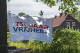 A banner displaying the text '75 JAAR VRIJHEID' is hanging outdoors, with a brick house and lush green trees in the background. The sky is clear with scattered clouds, suggesting a sunny day.