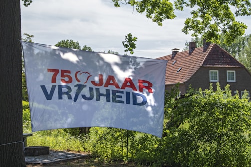 A banner displaying the text '75 JAAR VRIJHEID' is hanging outdoors, with a brick house and lush green trees in the background. The sky is clear with scattered clouds, suggesting a sunny day.