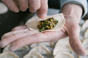 A close-up of hands preparing a dumpling, with a round dough wrapper being filled with a mixture of chopped vegetables and possibly meat. The hands are dusted with flour, and more dumplings can be seen in the background.