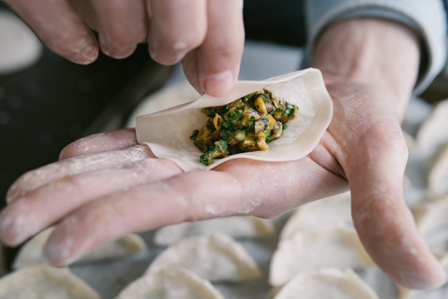 A close-up of hands preparing a dumpling, with a round dough wrapper being filled with a mixture of chopped vegetables and possibly meat. The hands are dusted with flour, and more dumplings can be seen in the background.