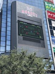 A tall building with a large electronic billboard displaying a video game screen, featuring a maze-like pattern and a score and timer at the top. The building is named 'MAGNET' and there are trees in the foreground.