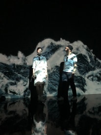 Two individuals stand against a dark backdrop with dynamic, wave-like patterns projected onto them. The lighting creates a surreal atmosphere as the patterns blend with their clothing, giving an impression of being immersed in an art installation.