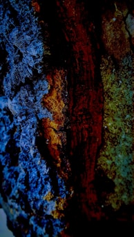 A close-up of a textured surface with rich colors, featuring a combination of deep blues, earthy browns, and subtle greens that create a visually striking contrast. The image appears to capture the intricate details of a natural material, possibly rust or a mineral deposit.