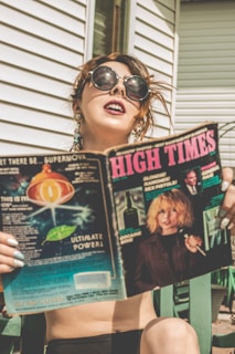 A person with large sunglasses and earrings is sitting outside next to a white building, holding a magazine titled 'High Times'. The magazine cover features imagery of cosmic elements and a person with blond hair holding a microphone.