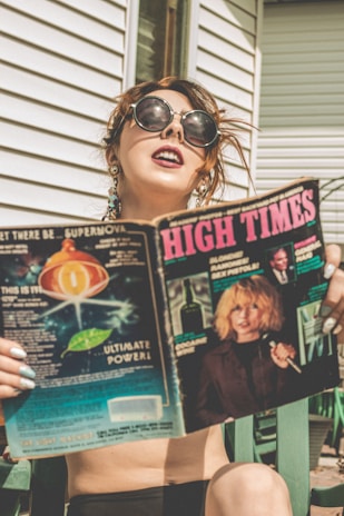 A person with large sunglasses and earrings is sitting outside next to a white building, holding a magazine titled 'High Times'. The magazine cover features imagery of cosmic elements and a person with blond hair holding a microphone.