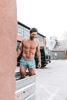 A muscular man standing in a metal tub filled with water, wearing colorful swim trunks. The setting appears to be an outdoor area with modern industrial buildings in the background and a parked black vehicle nearby. The man appears to be engaging in a cold water immersion activity.