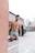 A muscular man standing in a metal tub filled with water, wearing colorful swim trunks. The setting appears to be an outdoor area with modern industrial buildings in the background and a parked black vehicle nearby. The man appears to be engaging in a cold water immersion activity.