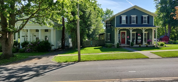Two houses are situated on a street lined with greenery. The house on the left is white with classical columns and surrounded by trees and shrubs. The house on the right is a two-story structure painted in dark blue, featuring white trim, a porch, and a bright red door. A road runs in the foreground with a clear sky and sunlight casting shadows.
