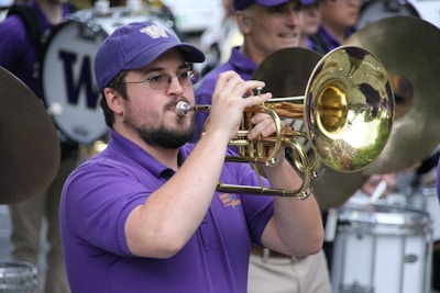 A musician in a purple shirt and cap plays a brass instrument in a marching band. The background shows other band members wearing similar attire, engaged with various percussion instruments.