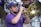 A musician in a purple shirt and cap plays a brass instrument in a marching band. The background shows other band members wearing similar attire, engaged with various percussion instruments.
