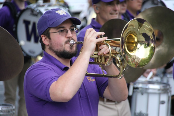 A musician in a purple shirt and cap plays a brass instrument in a marching band. The background shows other band members wearing similar attire, engaged with various percussion instruments.