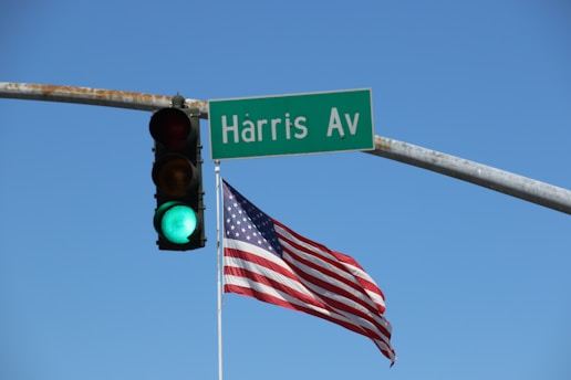 A street sign labeled 'Harris Av' is mounted on a metal pole, next to a traffic light that displays a green signal. An American flag is waving in the wind beneath the sign against a clear blue sky.