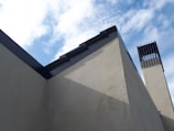 A modern architectural structure with clean, angular lines is set against a partly cloudy sky. The building features grey concrete walls and a prominent black metal trim along the roofline. To the right is a vertical, chimney-like structure with a louvered top section.