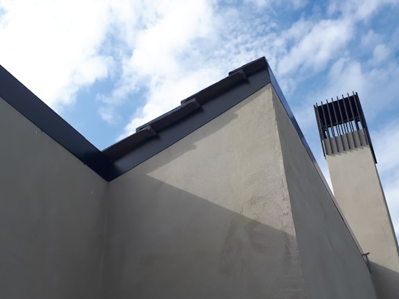 A modern architectural structure with clean, angular lines is set against a partly cloudy sky. The building features grey concrete walls and a prominent black metal trim along the roofline. To the right is a vertical, chimney-like structure with a louvered top section.
