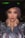 A model poses with hands elegantly placed around her neck, wearing a black wide-brimmed hat, a shimmering multicolored sequined top, and a pendant necklace. Her hair is styled in loose curls, and her makeup is bold with vibrant eye shadow and lipstick.