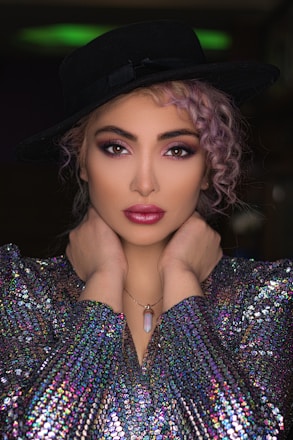 A model poses with hands elegantly placed around her neck, wearing a black wide-brimmed hat, a shimmering multicolored sequined top, and a pendant necklace. Her hair is styled in loose curls, and her makeup is bold with vibrant eye shadow and lipstick.