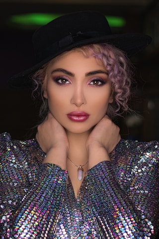A model poses with hands elegantly placed around her neck, wearing a black wide-brimmed hat, a shimmering multicolored sequined top, and a pendant necklace. Her hair is styled in loose curls, and her makeup is bold with vibrant eye shadow and lipstick.