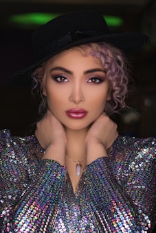 A model poses with hands elegantly placed around her neck, wearing a black wide-brimmed hat, a shimmering multicolored sequined top, and a pendant necklace. Her hair is styled in loose curls, and her makeup is bold with vibrant eye shadow and lipstick.
