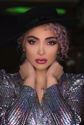 A model poses with hands elegantly placed around her neck, wearing a black wide-brimmed hat, a shimmering multicolored sequined top, and a pendant necklace. Her hair is styled in loose curls, and her makeup is bold with vibrant eye shadow and lipstick.