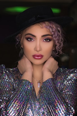A model poses with hands elegantly placed around her neck, wearing a black wide-brimmed hat, a shimmering multicolored sequined top, and a pendant necklace. Her hair is styled in loose curls, and her makeup is bold with vibrant eye shadow and lipstick.