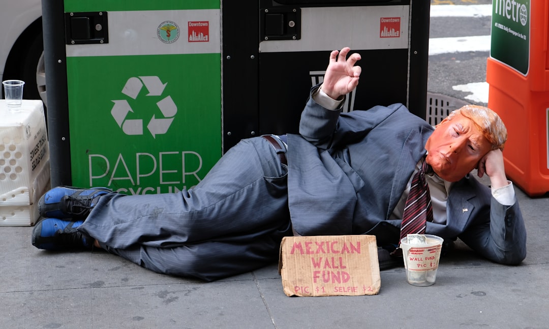 man in black jacket and blue denim jeans sitting on brown wooden seat, Donald Trump Street Performer