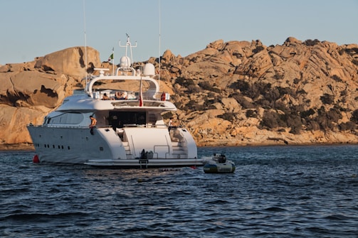 A large white yacht is anchored near a rocky coastline. The yacht has multiple decks, with visible people on the upper deck. The sun is low, casting a warm golden light on the rocks and water. A smaller inflatable boat is tethered to the yacht.