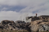 A person stands on rugged, rocky terrain, observing waves crashing against the rocks with a dramatic splash. The sky is overcast with clouds, adding to the sense of adventure and exploration.