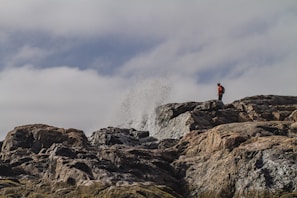 A person stands on rugged, rocky terrain, observing waves crashing against the rocks with a dramatic splash. The sky is overcast with clouds, adding to the sense of adventure and exploration.