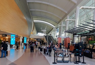 A modern airport terminal features a high, arched ceiling with large windows on one side. People are walking or seated in the area, some holding luggage. Bright blue digital signs mark baggage claim areas. A Starbucks coffee shop is located on the right, with several customers lined up. An escalator is visible in the center, leading to an upper level.